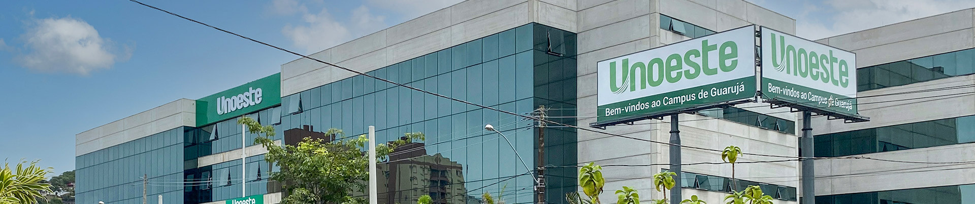 Edifício moderno do Unoeste Campus Guarujá, com uma fachada de vidro espelhada e placas de sinalização da universidade. Um outdoor proeminente dá as boas-vindas ao campus de Guarujá, destacando a presença da universidade no litoral paulista. Esta unidade oferece graduação e diversos cursos superiores, reforçando a educação de qualidade na região.