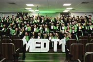 Grupo de formandos do curso de medicina da Unoeste, celebrando com becas e faixas verdes. Eles estão reunidos em um auditório, posando em frente às letras grandes que formam "MED T1".