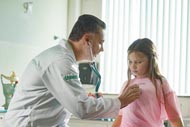 Médico da Unoeste (Universidade do Oeste Paulista) em consulta, utilizando um estetoscópio para examinar uma criança que veste uma camiseta rosa. Eles estão em um consultório médico iluminado pela luz natural que entra pela janela.