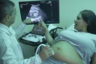 Médico fazendo exame de ultrassom na barriga de uma grávida, com a gestante olhando para a imagem do bebê na tela.